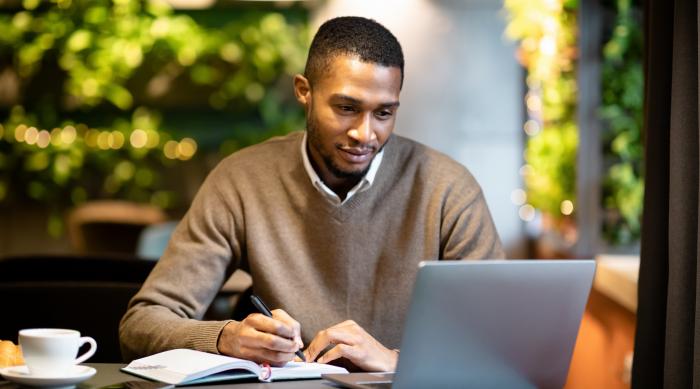African American man writing in notebook sitting looking at laptop